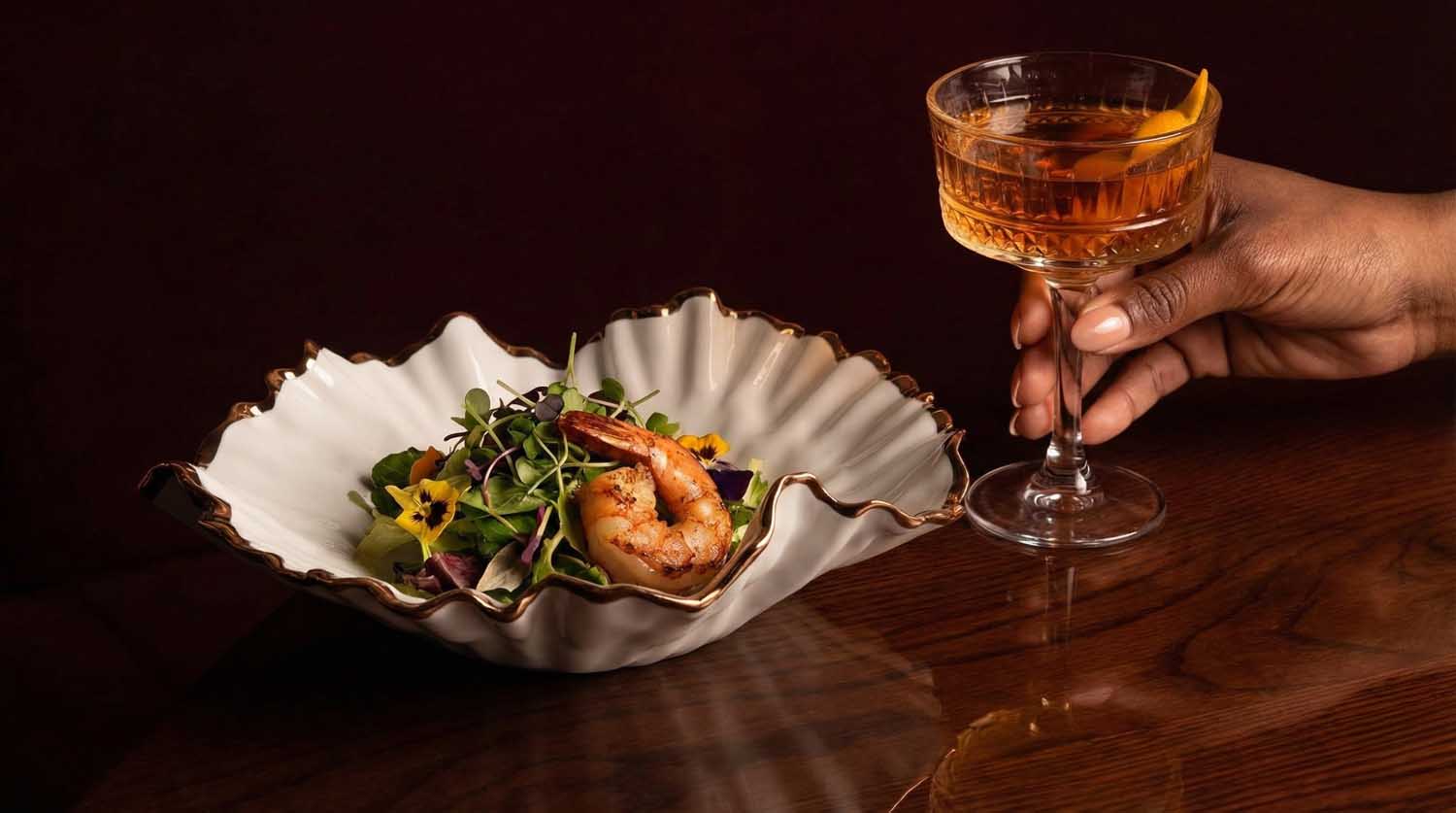 Ruffled porcelain bowl with prawn salad beside a hand holding a cocktail, custom restaurant plate